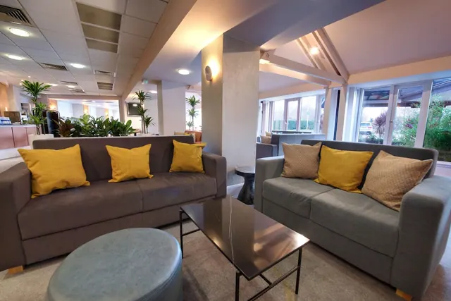 A lounge seating area in a hotel lobby with two sofas, yellow cushions, a coffee table and a leather pouf, with windows looking out onto greenery.