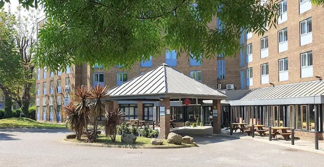 The brick front of a hotel with a metal-roofed entrance canopy, a tree in the foreground, picnic benches on a covered terrace to the right, and a paved drive.