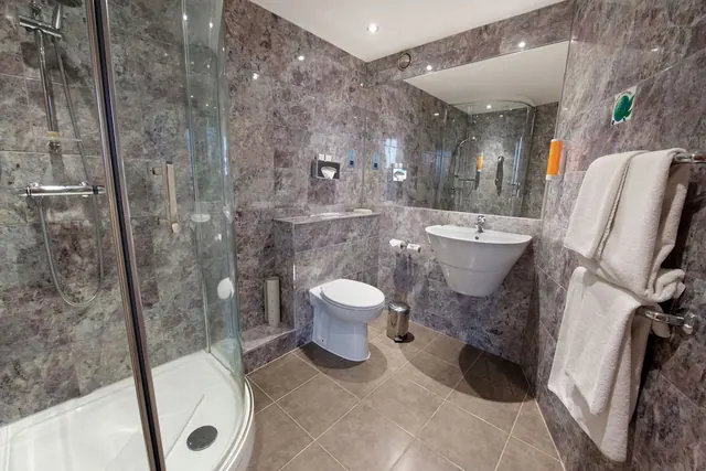 A bathroom with grey marble-effect tiles, a corner glass shower cubicle, a wall-mounted basin, a toilet and white towels on a rail.