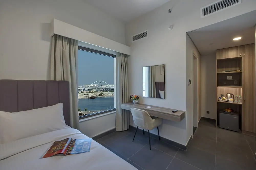 A bedroom with a made bed, a wall-mounted desk and chair, a kettle and small fridge by the wardrobe, and a window looking over a waterway with a white arch bridge.