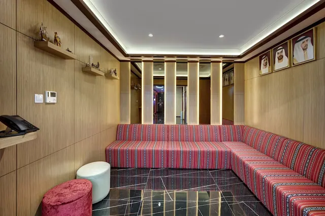 A lounge seating area with a red striped built-in banquette along the right wall, wood-panelled walls, a dark marble floor, framed portraits and two ottomans.