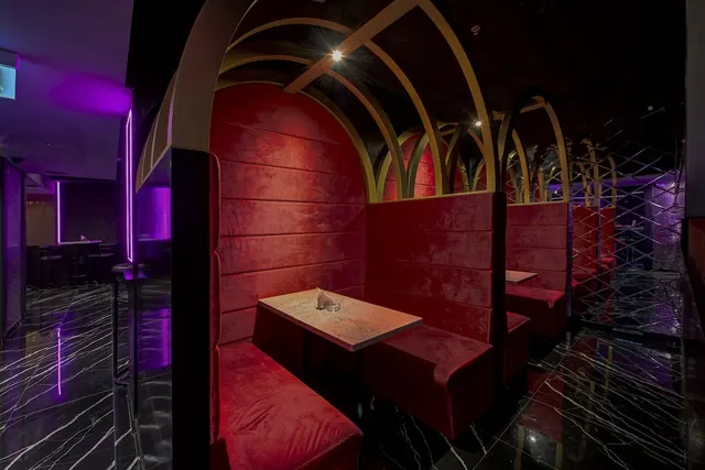 A dimly lit lounge with red velvet arched booth seating, a marble-topped table, and purple neon lighting along the walls.