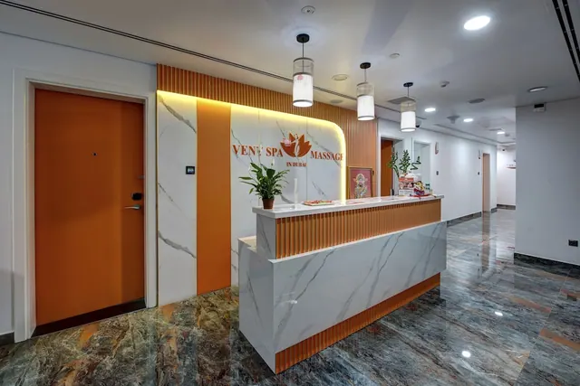 A spa reception counter with a white marble front and orange slatted panels stands beneath a backlit wall sign reading Vent Spa Massage, with an orange door on the left and pendant lights overhead.