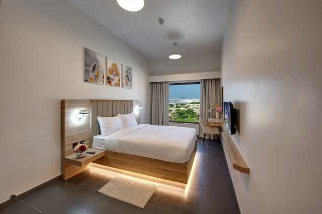 A double bed with white bedding sits beside a wall-mounted television and a small desk, with a window showing green trees and a lawn outside.