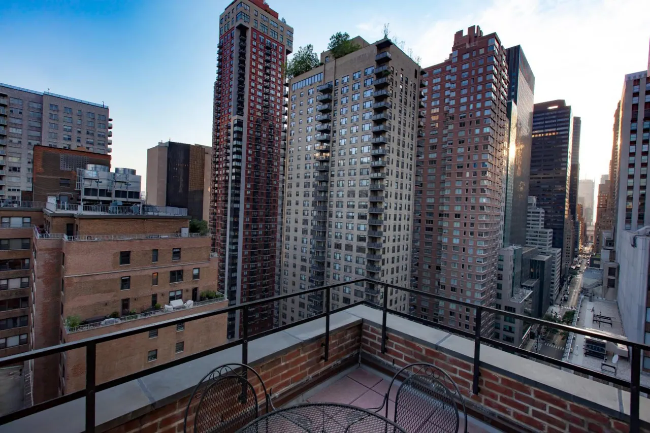 A brick-walled terrace with a metal railing and two wrought-iron chairs at a small table overlooks tall residential towers and midrise buildings.