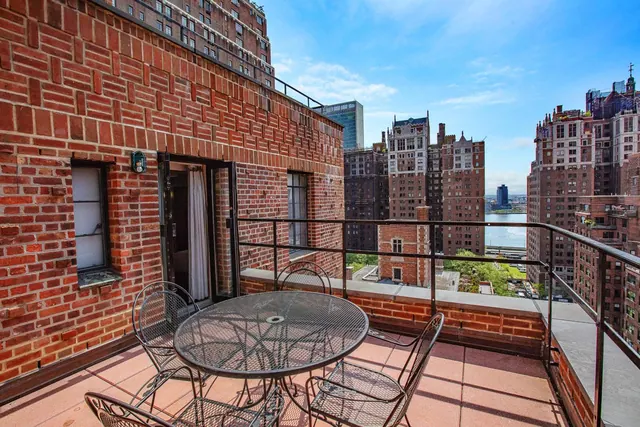 A brick-walled terrace with a metal table and four chairs overlooks high-rise buildings and a river under a blue sky.