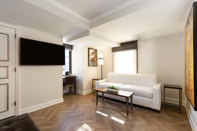 A living area with a wall-mounted television, a white sofa, a wooden coffee table and a desk with a chair near the window.