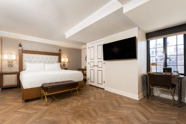 A bedroom with a king bed, an upholstered bench, a wall-mounted television, double closet doors and a desk by a window overlooking city buildings.