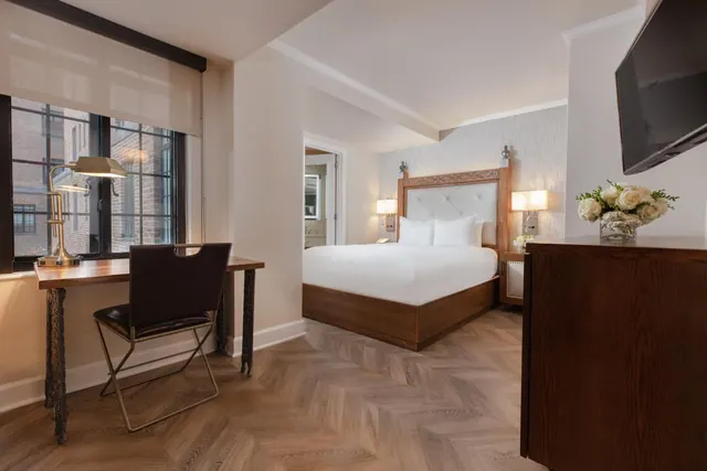 A bedroom with a king bed, wooden headboard, wall-mounted television, and a desk by a window looking onto a brick building.