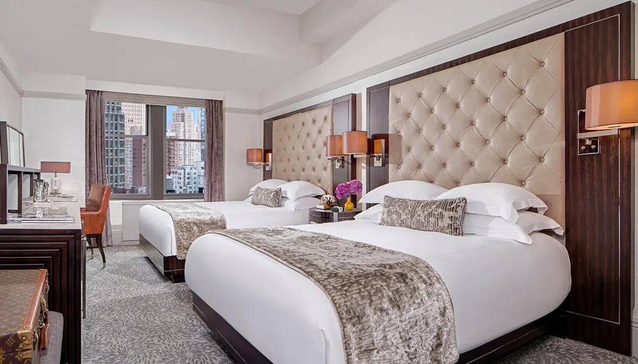 A bedroom with two double beds with tufted upholstered headboards, a desk on the left, and a window showing high-rise buildings.