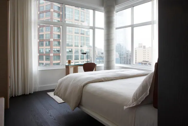 A bedroom with a made bed with white linen, a small desk and chair, and corner windows looking out over city buildings.