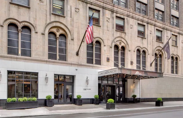 The stone facade of the hotel with arched windows, an American flag, and a canopied entrance with potted shrubs on the sidewalk.