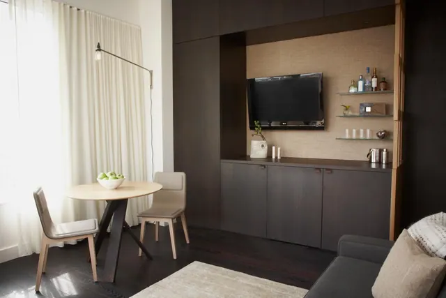 A living area with a round wooden dining table and two upholstered chairs, a wall-mounted television set into a dark wood cabinet with bar shelves, and a grey sofa at the right.