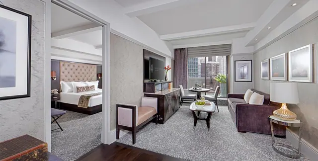 A hotel suite living area with a sofa, coffee table, wall-mounted television and a glimpse of a bedroom with a bed through the doorway, with city buildings visible through the glass doors.