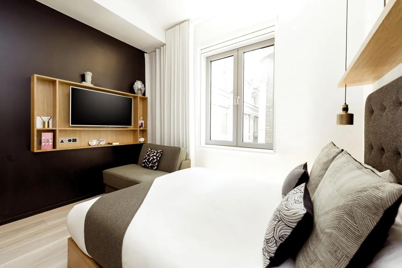 A bedroom with a double bed with patterned cushions, a wall-mounted television on a wooden shelf unit, a upholstered bench seat, and a window looking onto a neighbouring building.