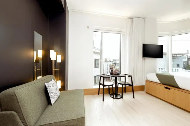 A hotel room with a grey sofa, a small round table with two chairs by a window, and a wall-mounted television beside a bed.