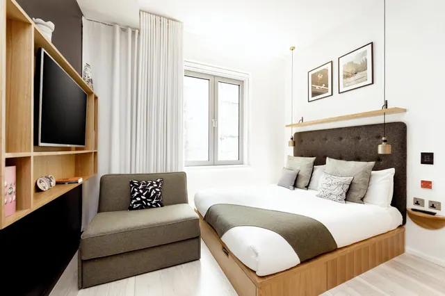 A small hotel bedroom with a double bed, a grey upholstered armchair, a wall-mounted television on open wooden shelving, and a window with white curtains.