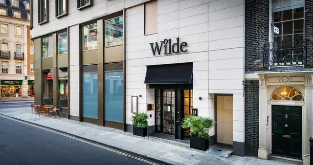 Street-level view of the Wilde hotel entrance with a black canopy, potted palms, and a neighbouring brick townhouse with a dark green door.