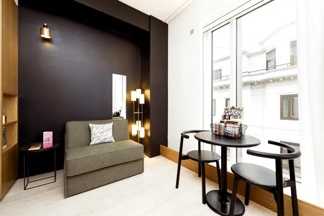 A small room with a grey sofa bed against a dark accent wall, a round black table with two chairs, and a tall window looking onto a neighbouring white building.