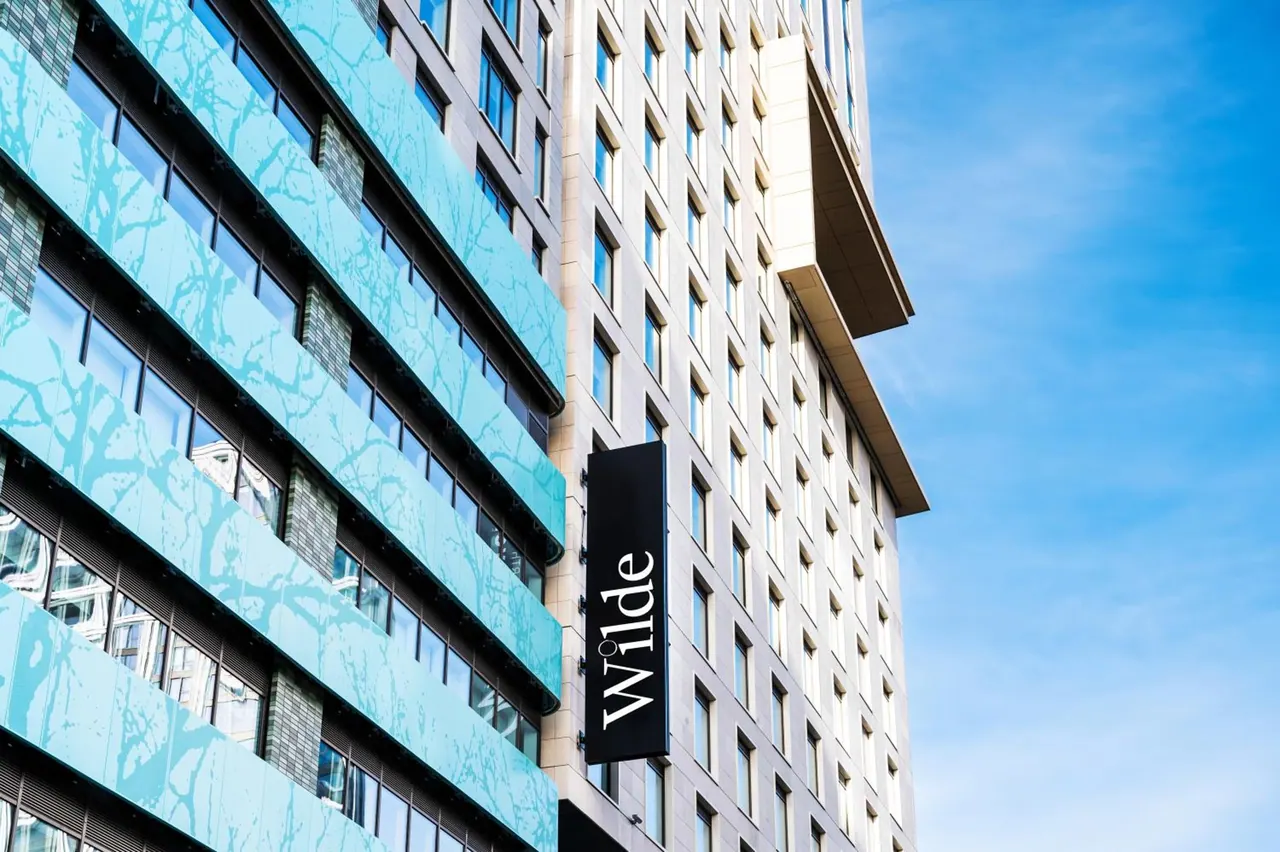 The exterior of a tall building with teal patterned panels and a vertical black sign reading 'wilde' against a clear blue sky.
