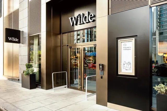The street entrance of the Wilde hotel, with a dark metal facade, a lit Wilde sign above glass doors marked Welcome, and a wall panel reading Welcome to Paddington.