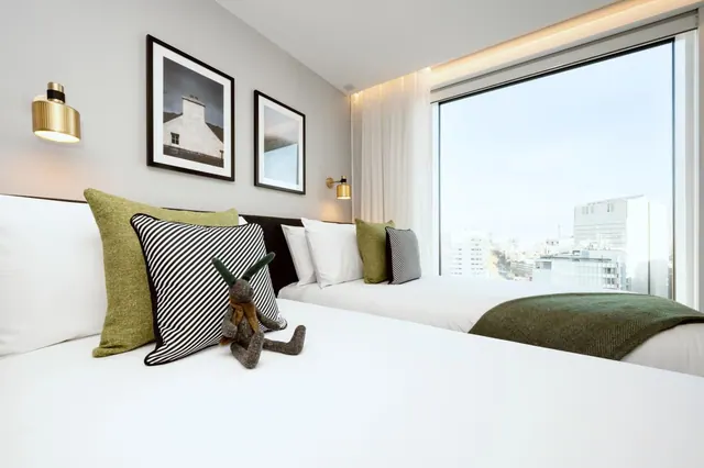A bedroom with two beds dressed in white linen and green cushions, framed black-and-white prints on the wall, and a large window overlooking a city skyline.