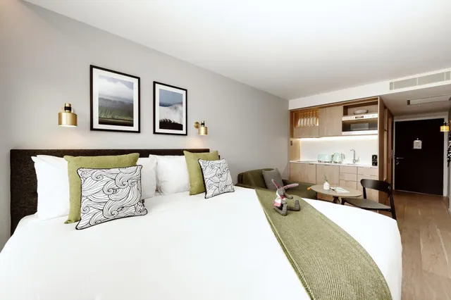 A made king bed with green and patterned cushions sits in a bedroom beside a wooden kitchenette, a kettle and a green sofa.