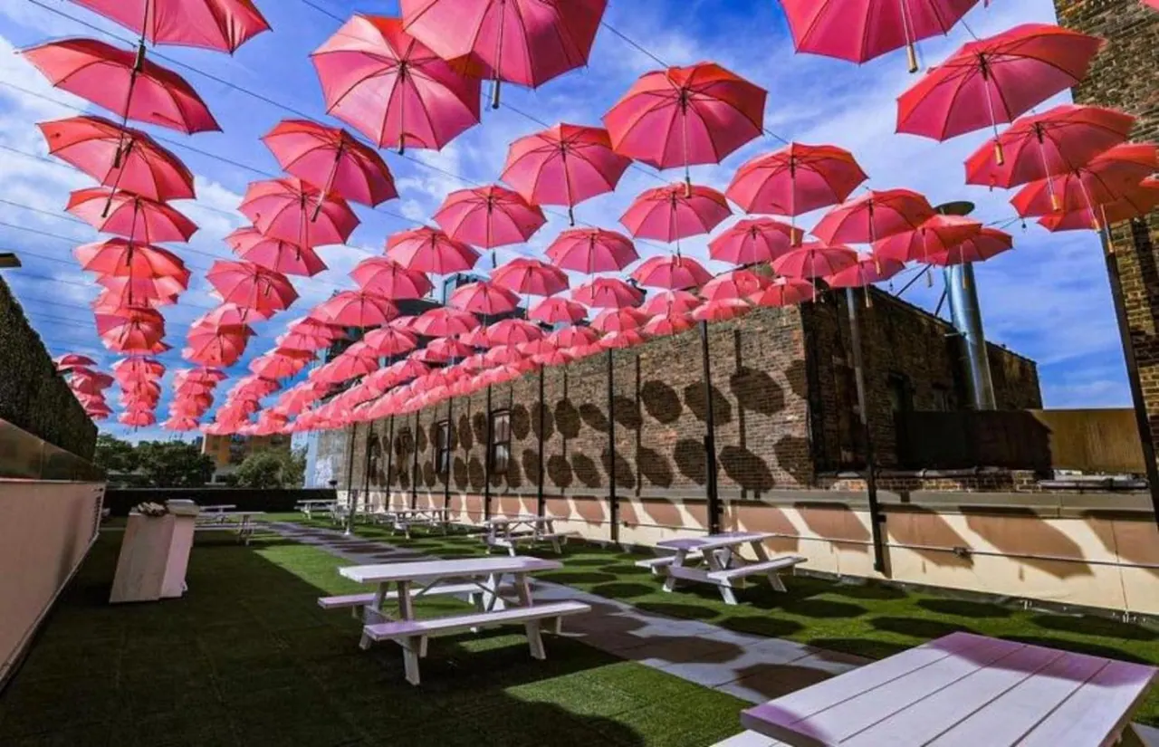 A rooftop terrace with rows of pink umbrellas hung overhead, artificial turf, and white picnic tables beside a brick building wall.