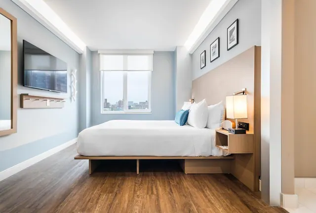 A bedroom with a made king bed, a wall-mounted television, and a window showing low-rise city buildings in the distance.