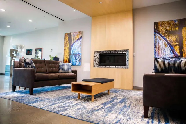 A lounge seating area with a brown sofa, a brown armchair, a wooden coffee table, a wood-panelled fireplace and large abstract paintings on a blue rug.