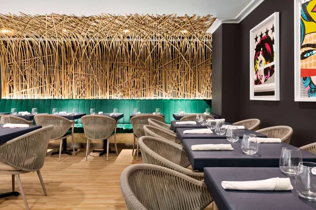 A restaurant dining room with a woven bamboo ceiling installation, green banquette seating, navy tables set with folded white napkins and glasses, rope-backed chairs and pop art prints on the wall.