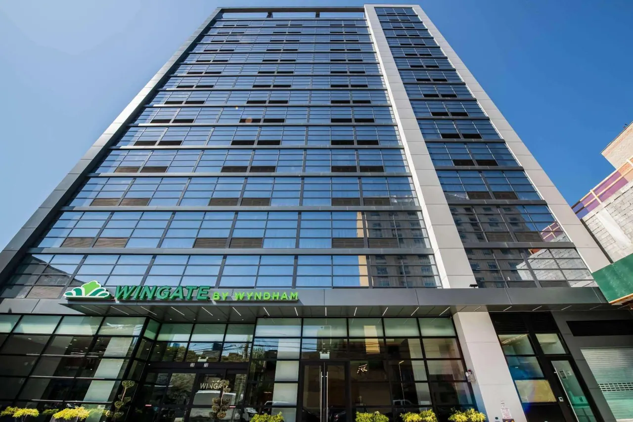 Low-angle view of a tall glass hotel tower with a Wingate by Wyndham sign above the entrance under a clear blue sky.