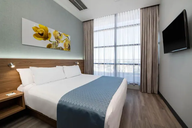 A bedroom with a made king bed, a wall-mounted flat-screen television and floor-to-ceiling windows with sheer curtains looking out over city buildings.