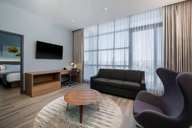 A living area with a dark grey sofa, a round wooden coffee table on a patterned rug, a wall-mounted television and a desk beside floor-to-ceiling windows, with a bed visible through the doorway on the left.