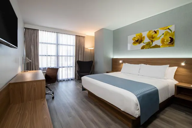 A hotel bedroom with a king bed, a wall-mounted television, a wooden desk with a chair, an armchair and large windows with sheer curtains.