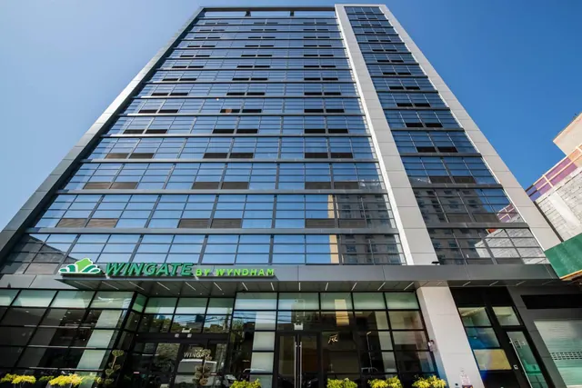A low-angle view of a tall glass-fronted hotel tower with a Wingate by Wyndham sign above the entrance under a clear blue sky.
