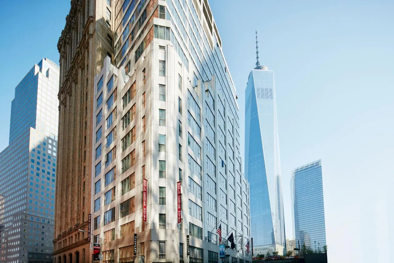 Exterior of a tall hotel building with vertical red banners on its facade, with One World Trade Center rising in the background.