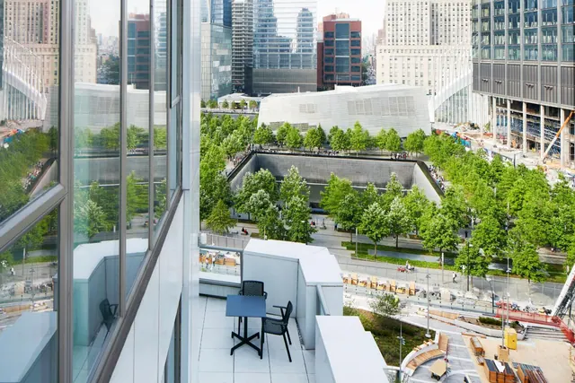 A white balcony with a dark table and two chairs overlooks a sunken memorial pool, rows of trees, a plaza, and city towers.