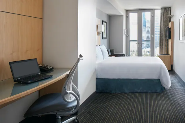 A hotel bedroom with a laptop on a curved desk and chair, a bed with white bedding, a seating chair, and tall buildings visible through glass doors.