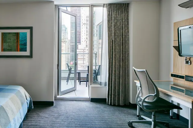 A hotel room with a bed, a desk with a chair and wall-mounted television, and an open door to a balcony overlooking city high-rises.