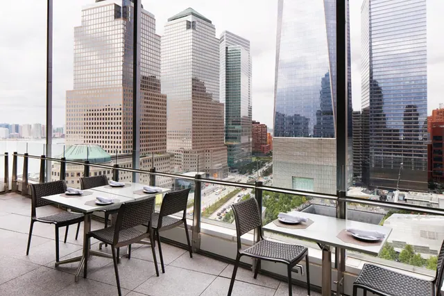 An outdoor terrace with set dining tables and chairs overlooks Lower Manhattan skyscrapers, including One World Trade Center, with water visible at the left.