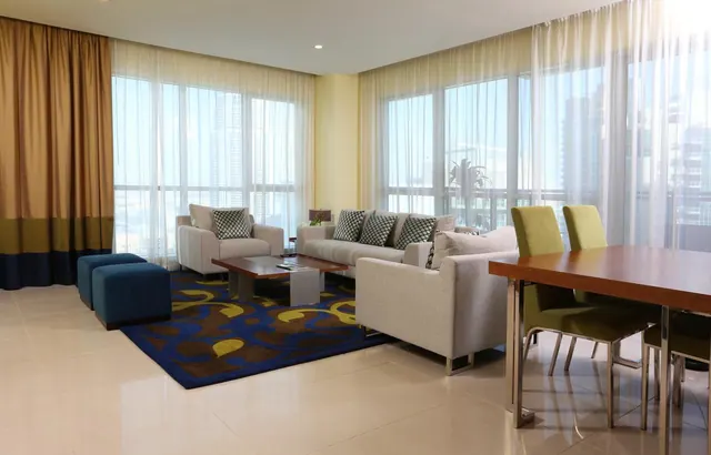 A living area with grey sofas and armchairs around a wooden coffee table on a blue patterned rug, with a dining table and green chairs at right and tall windows showing city towers.
