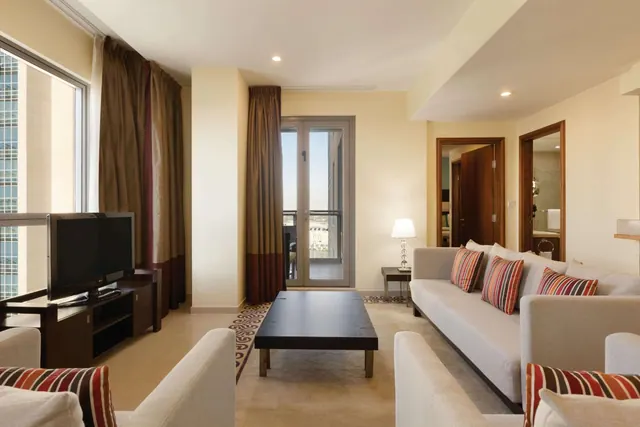 A living room with a grey sofa, armchairs, a low coffee table, a flat-screen TV and a glass balcony door showing buildings outside.
