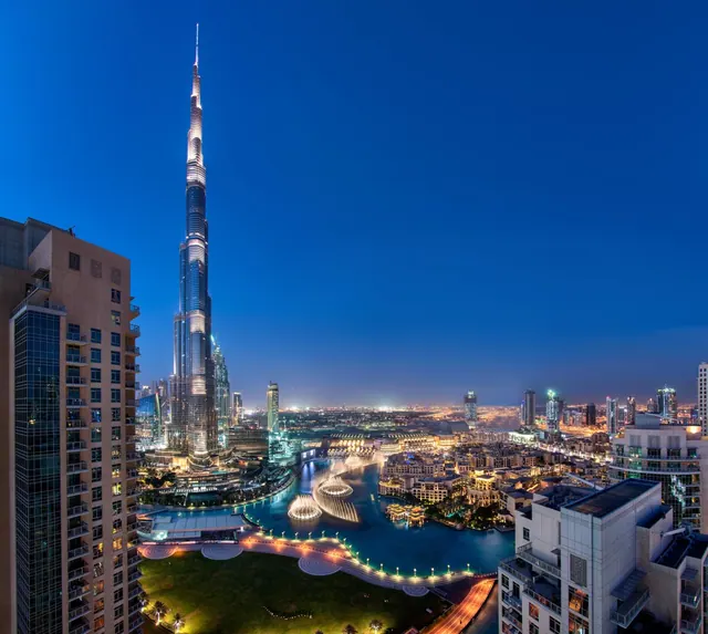 Elevated night view of the Burj Khalifa tower, an illuminated fountain lake and surrounding city buildings under a deep blue dusk sky.