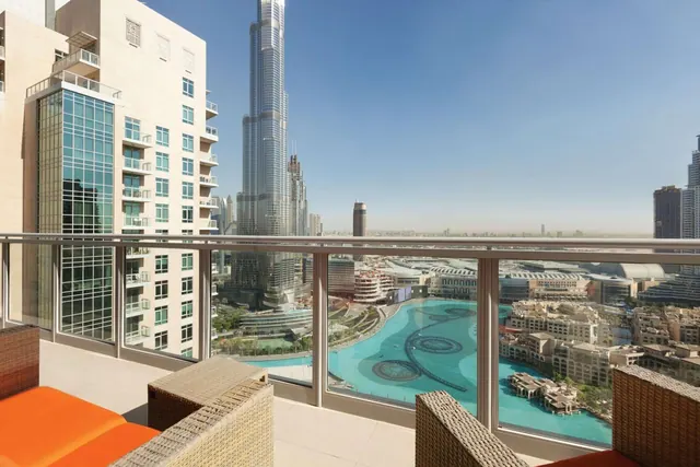 A balcony with wicker seating and orange cushions overlooks the Burj Khalifa tower, a turquoise lake and surrounding city buildings.
