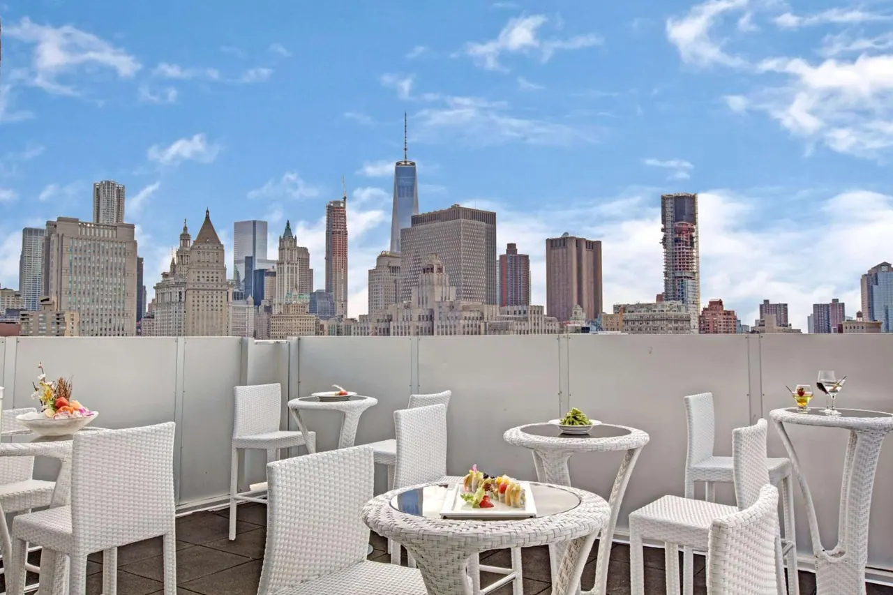 A rooftop terrace with white wicker tables and chairs, with a city skyline including One World Trade Center behind a white wall.