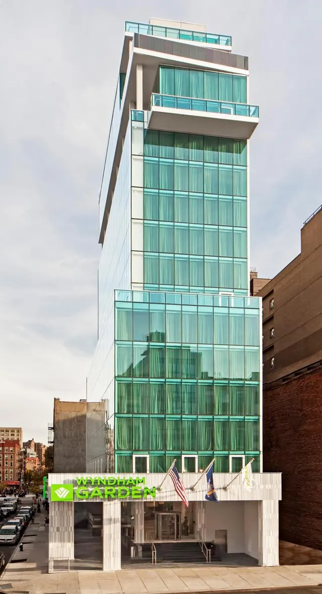 Exterior of a tall glass-fronted hotel tower with green Wyndham Garden signage above the entrance on a city street.