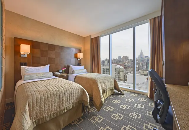 A twin-bedded hotel room with floor-to-ceiling windows showing city buildings and the Empire State Building in the distance.