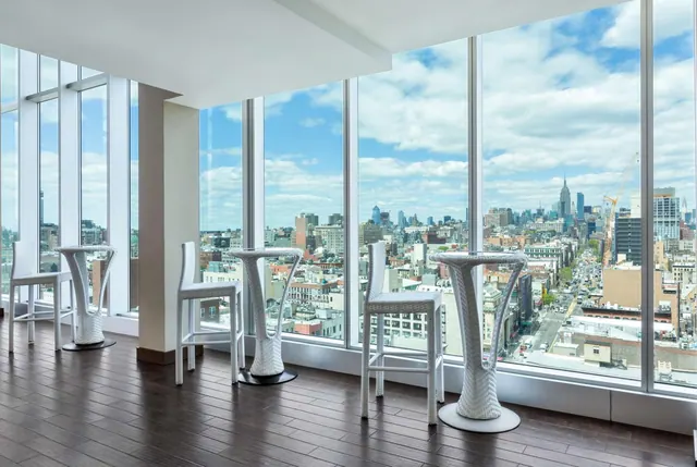 A lounge area with white wicker high tables and stools sits beside floor-to-ceiling windows overlooking a city skyline that includes the Empire State Building.