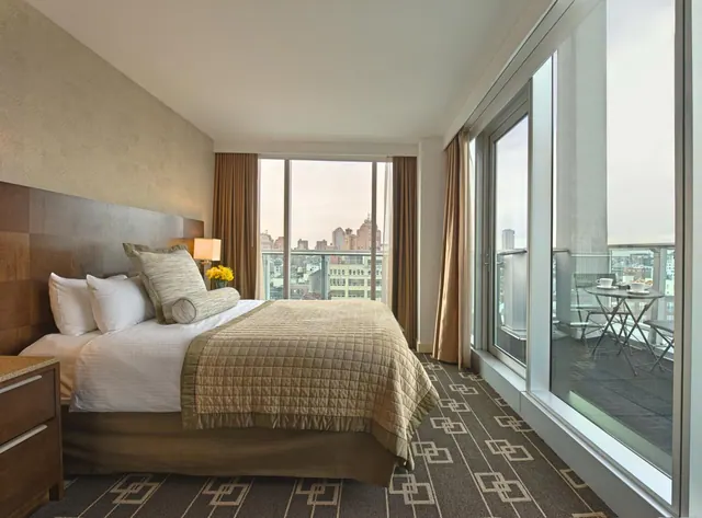 A bedroom with a made bed, floor-to-ceiling windows and a private balcony with a table and chairs, with city buildings visible beyond.